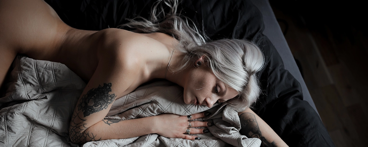 WitheredRose silberblond Rückansicht auf dunklem Bett mit großem Rose-Blumen-Schulter-Tattoo – Querformat