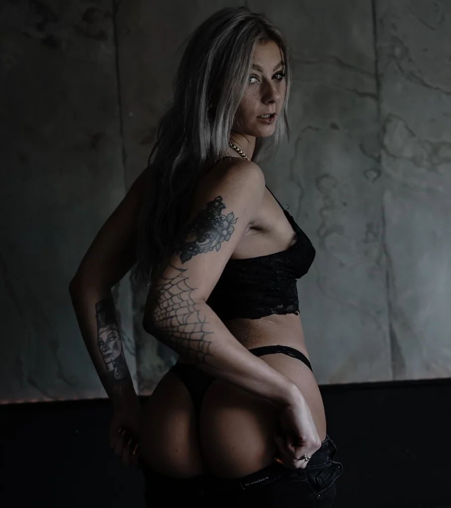 WitheredRose silberblond im schwarzen Spitzen-BH mit offener Jeans und Po sichtbar – Spinnennetz-Sleeve mit Morticia-Portrait-Tattoo